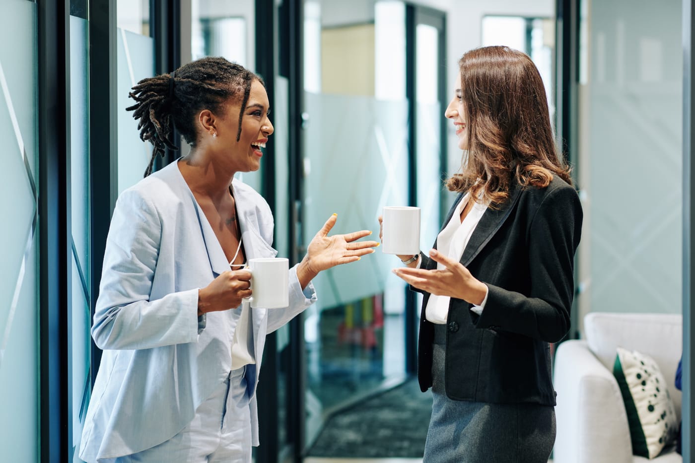 Deux femmes souriantes discutent et gesticulent en tenant des tasses dans un bureau moderne