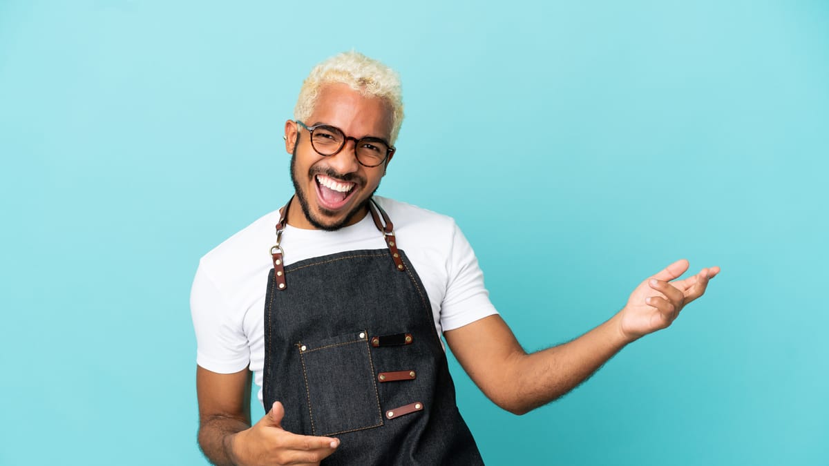 Homme souriant et barbu, cheveux blonds, lunettes, tablier noir, gestuelle ouverte, fond bleu clair