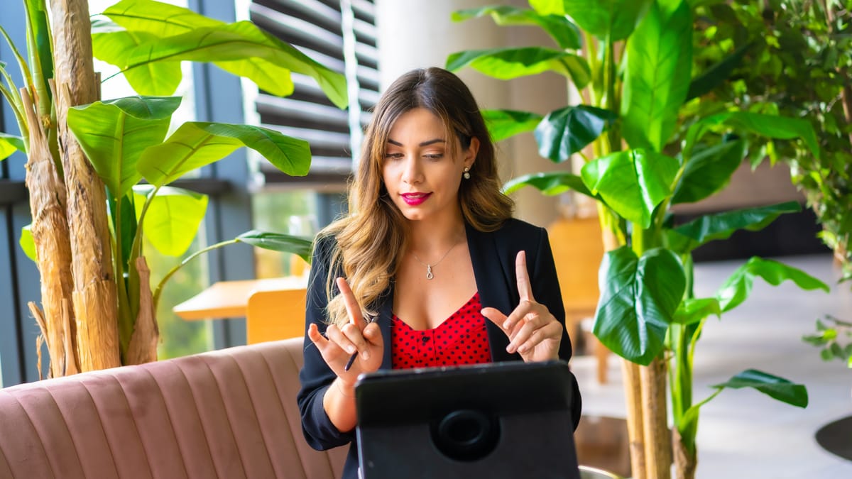 Femme en blazer noir et haut rouge, utilisant une tablette, faisant des gestes explicatifs dans un bureau moderne avec plantes