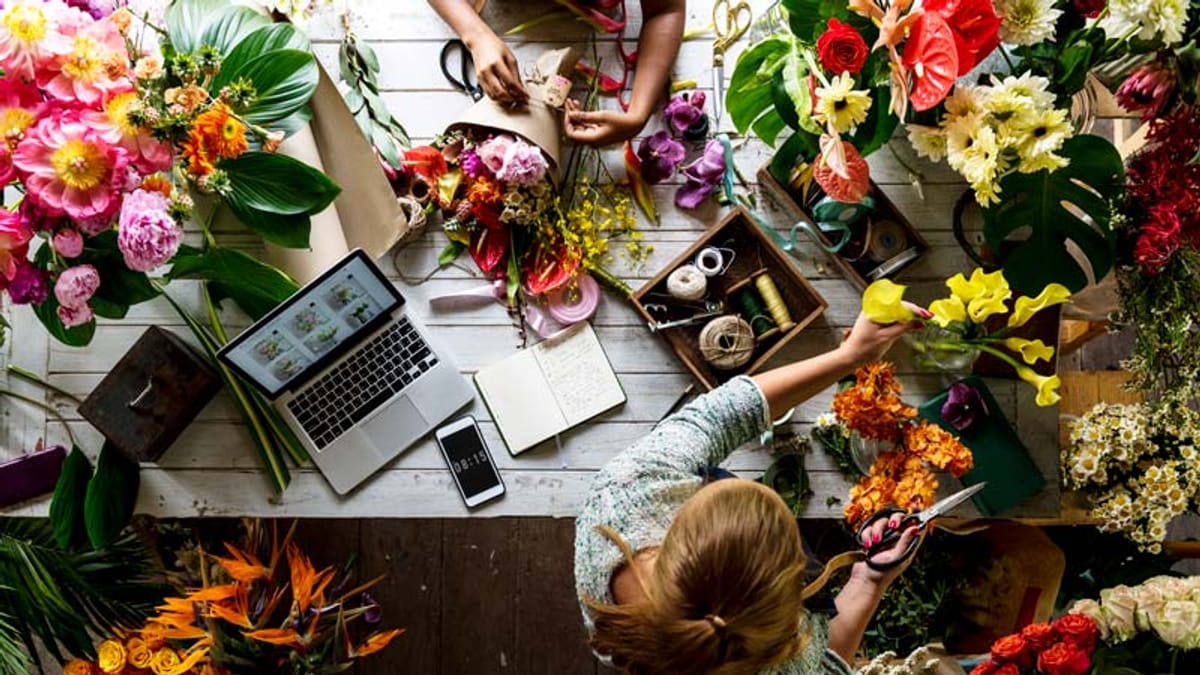 Deux fleuristes travaillent sur une table remplie de fleurs colorées, d'outils, un ordinateur portable et un smartphone